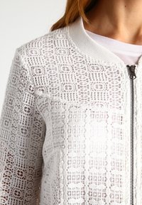 Bílá bomber bunda vyrobená z krajkového materiálu, s zipovým zapínáním, žebrovaným límcem a složitými geometrickými vzory po celém povrchu.
