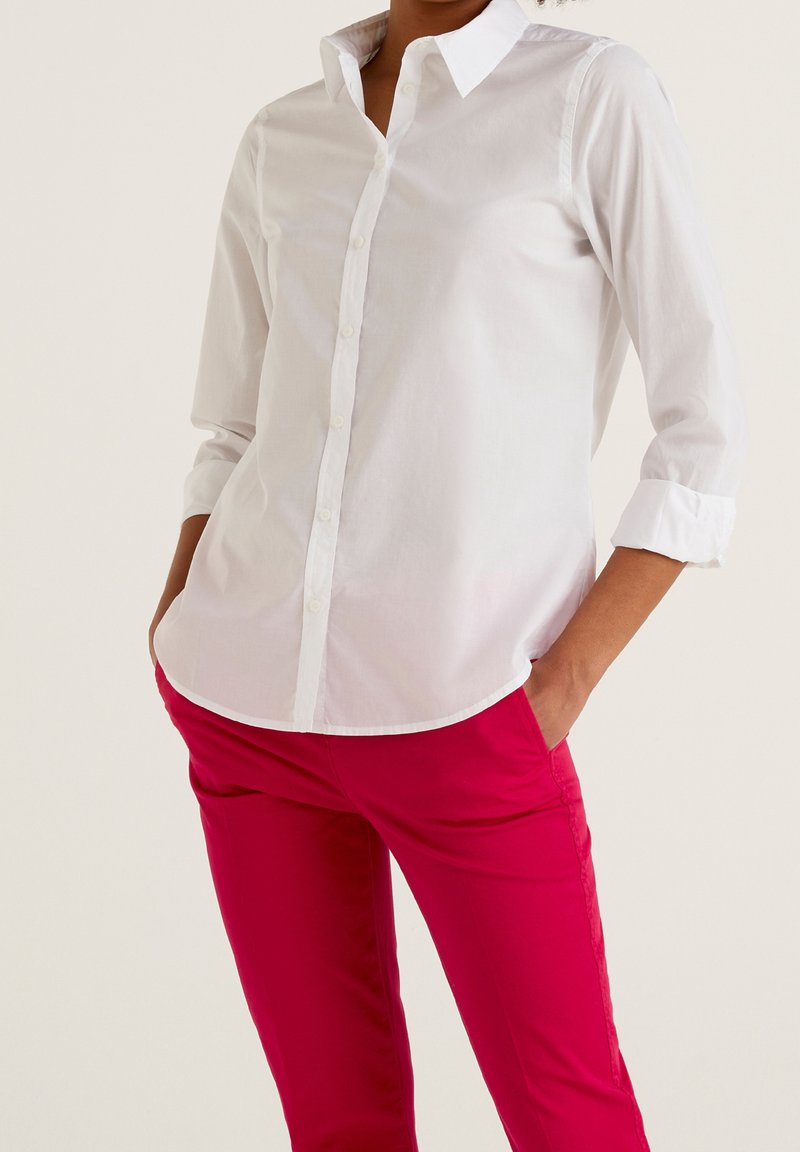 Camisa blanca de botones con mangas largas, combinada con pantalones de color rosa brillante. La camisa tiene un cuello y las mangas enrolladas, fabricada en material de algodón.