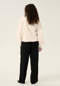 Ljusbeige stickad tröja med ett texturerat mönster på ryggen, i kombination med lösa svarta byxor. Tyget verkar mjukt med ballongärmar.