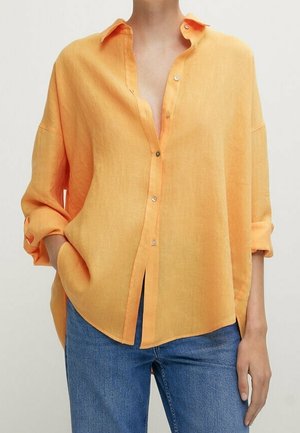 Loose-fitting, long-sleeve orange button-up shirt with rolled cuffs paired with blue denim jeans, hands in pockets, on neutral background.