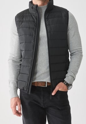 Uomo che indossa un gilet trapuntato nero sopra a un maglione grigio chiaro, pantaloni neri, cintura marrone e orologio da polso argentato con una mano in tasca.
