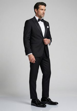 Man die zijn zwarte smokingjasje verstelt, met een wit overhemd, zwarte vlinderdas, zwarte nette broek en gepoetste zwarte nette schoenen, staand op een grijze achtergrond.