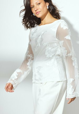 Hvid gennemsigtig bluse med lange ærmer, prydet med broderede blomsterdesigns og rund halsudskæring. Sammen med cremefarvede bukser.