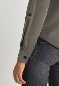 Left arm wearing an olive green long-sleeve shirt with buttoned cuff, resting beside a hip in black denim jeans with back pocket visible.