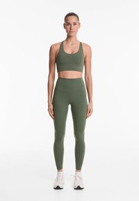 Olivnozeleni športni modrček in hlače z visokim pasom iz gladkega, raztegljivega materiala. Model stoji obrnjena naprej v športnih čevljih.