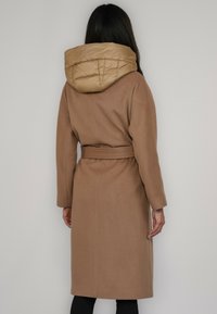 Długi brązowy płaszcz z paskiem w talii i dużym, puchowym kapturem w kolorze beżowym. Posiada długie rękawy i prosty krój sięgający do połowy łydki.