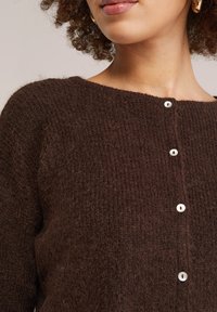 Sieviete valkā mīkstu, tumši brūnu adītu kardiganu ar baltām pogām un zelta riņķveida auskariem, redzams kakla izgriezums un augšējā torss.