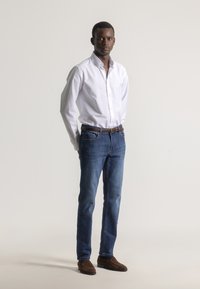 Camisa blanca abotonada, vaqueros azules de mezclilla, cinturón marrón y zapatos de ante. Ajuste ceñido, textura suave, diseño clásico. De pie contra un fondo liso.