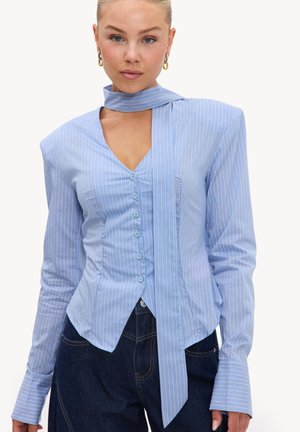 Femme portant un chemisier ajusté rayé bleu clair avec des manches longues, des poignets allongés, une cravate attachée au cou et un jean bleu foncé.