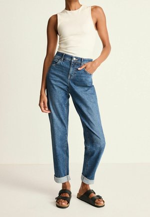 Kobieta w białym topie bez rękawów, wysokim stanem niebieskich dżinsów z podwiniętymi nogawkami oraz czarnych sandałach na klamrę, stoi z jedną ręką w kieszeni.