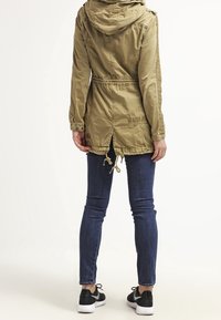 Veste à capuche vert olive avec taille à cordon, poches latérales et accents texturés. Portée avec un jean foncé et des baskets noires.