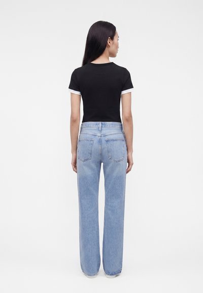 Γυναίκα με μακριά σκούρα μαλλιά στέκεται με την πλάτη στραμμένη, φοράει μαύρο κοντομάνικο μπλουζάκι και ανοιχτό μπλε τζιν με ψηλή μέση σε λευκό φόντο.