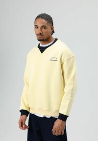 Gele trui met een V-hals, zwarte accenten op de kraag en manchetten, met de tekst "STUDIO ABYSS" in het zwart. Zachte stof, ontspannen pasvorm.