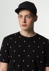 Sort bomulds t-shirt med hvide timeglasmønstre og en rund halsudskæring, parret med en sort struktureret kasket med en buet skygge.