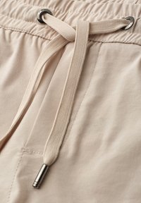 Pantalon en tissu beige avec un cordon plat noué passant à travers des œillets métalliques à la taille élastique.