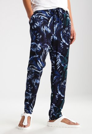 Persoon die een donkerblauwe broek met tropische bladerenprint, witte sandalen en een wit overhemd draagt, staand met de handen in de zakken.