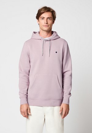 Junger Mann trägt einen hellvioletten Hoodie mit Fronttasche und weiße Hose, steht vor einem schlichten hellen Hintergrund.
