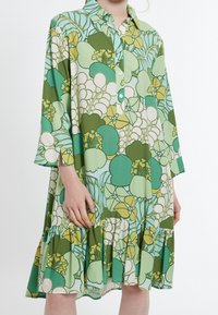 Grön blommönstrad skjortklänning med krage, knappstängning framtill, trekvartslånga ärmar och en volangnedre del. Har stora, stiliserade blommönster.