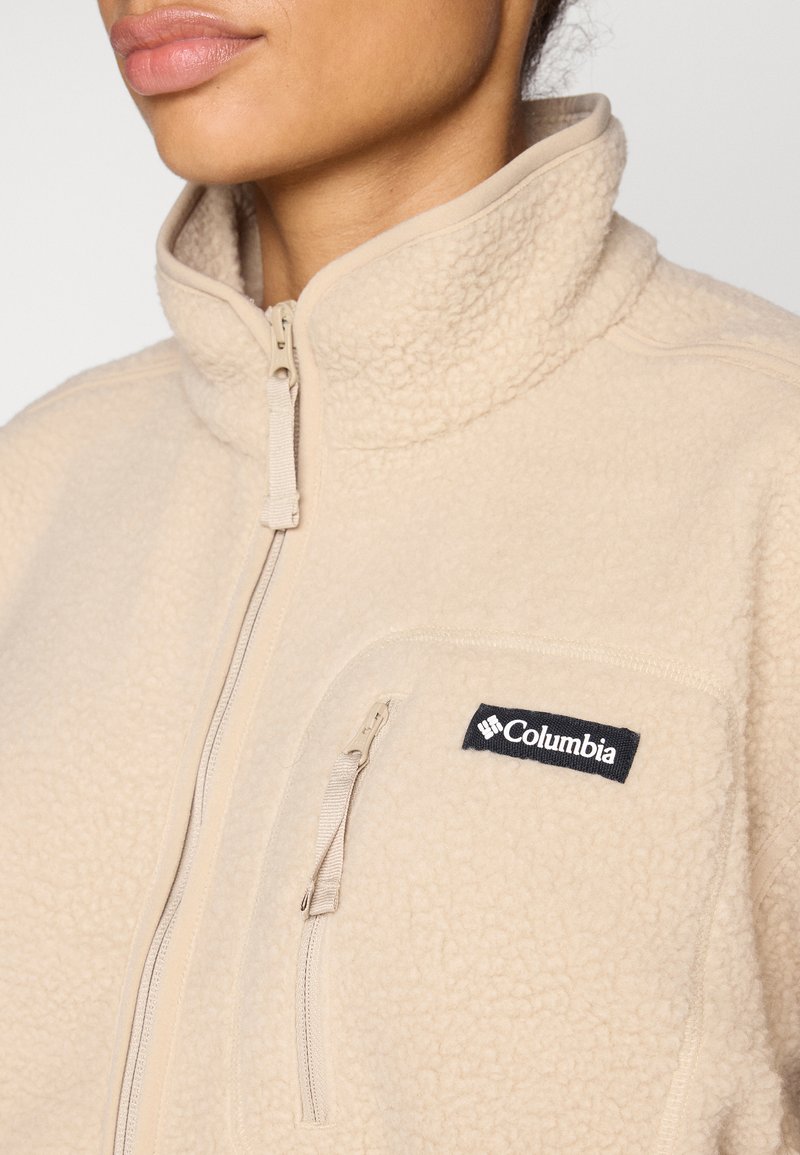 Nahaufnahme einer Person, die eine beige Columbia Fleecejacke mit Reißverschlusstasche auf der Brust und hohem Kragen trägt.