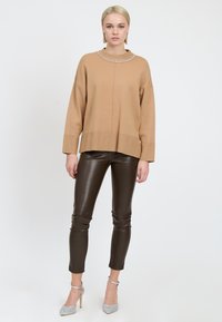 Maglione beige lavorato a maglia con scollo tondo decorato con strass, abbinato a pantaloni slim in ecopelle marrone e scarpe argentate con tacco alto.