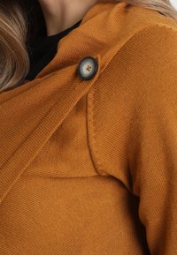 Mosterdgele gebreide cardigan met een wikkeldesign, een grote zwarte knoopaccent en een gestructureerde stof.