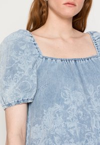 Top de denim azul claro com mangas curtas bufantes, decote quadrado e padrão floral. Apresenta um acabamento elástico e detalhes de bolso lateral.