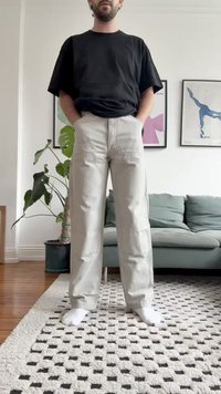 Calças cargo cinza claro com bolsos frontais e detalhes em costura, combinadas com uma t-shirt preta. A pessoa está de pé sobre um tapete texturizado, com os pés descalços visíveis.
