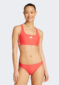 Koralli bikini-setti, jossa on avara kaula-aukko ja valkoinen adidas-logo, sekä samalla korallinsävyiset matalavyötäröiset alaosat, valmistettu sileästä joustavasta kankaasta.