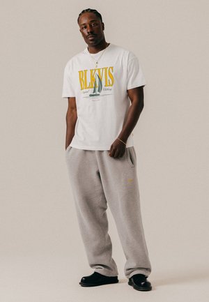 Homme aux cheveux tressés portant un t-shirt graphique blanc, un pantalon de survêtement gris, des chaussures noires, un collier en or et un bracelet sur un fond uni.