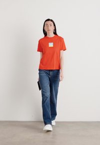 Rode katoenen t-shirt met een vierkante graphic op de borst, gecombineerd met blauwe jeans en witte sneakers, vastgehouden met een zwarte clutch. Soepele textuur, casual pasvorm.