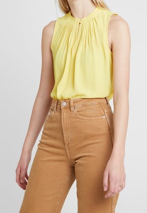 Mujer con blusa amarilla sin mangas y plisada, metida dentro de unos vaqueros de tiro alto de color marrón claro, de pie contra un fondo liso.