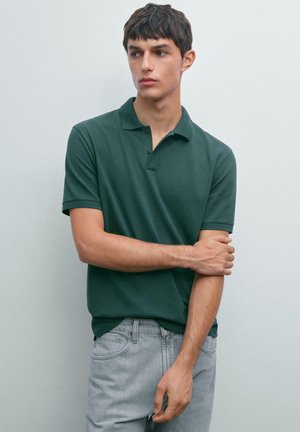 Ung man som bär en mörkgrön kortärmad polo och ljusgrå jeans, stående mot en enfärgad ljusgrå vägg, med blicken riktad åt sidan.