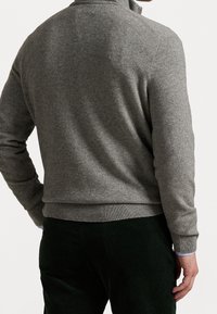 Pull gris à col montant, avec poignets et ourlet côtelés, en tissu doux et texturé, porté sur un pantalon en velours côtelé vert foncé.