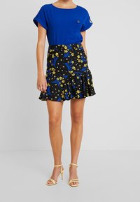 Top blu a maniche corte con tasca, abbinato a una gonna nera con motivi floreali gialli e accenti blu, rifinita con un orlo arricciato.