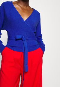 Person in einem blauen Wickelstrickjacke mit gebundenem Gürtel und leuchtend roten hochgeschnittenen Hosen, die die Hände in den Taschen hat, vor einem hellen Hintergrund.