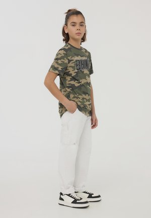 T-shirt w kolorze kamuflażu zielonym z czarnym nadrukiem, w połączeniu z białymi dżinsami i czarnymi trampkami. T-shirt ma krótkie rękawy i okrągły dekolt.