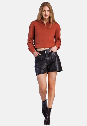 Ung kvinde iført en rustfarvet cropped sweatshirt, sorte lædershorts og sorte ankelstøvler, stående med hænderne i lommerne.