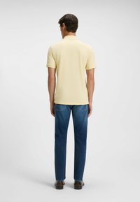 Man som bär en blekgul polotröja, blå jeans och mörkbruna skor, står vänd bortåt mot en jämn ljus bakgrund.