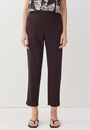 Femme portant un pantalon droit marron foncé coupé au-dessus de la cheville, associé à un haut sans manches noir et blanc à motifs, et des sandales noires.