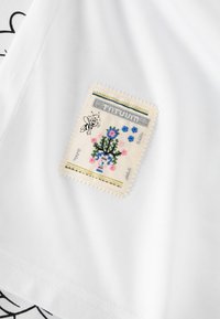 Hvid T-shirt med et stofmærke med broderede blomster, en blå vase, en bi og bølgende kanter.