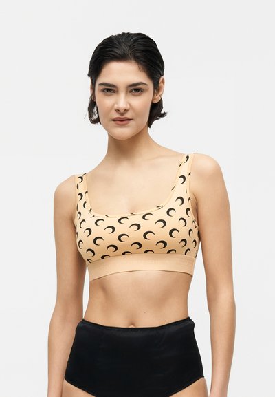 Mulher com cabelo curto e escuro, vestindo um top cropped bege com padrão de lua crescente preta e calções pretos de cintura alta, em pé contra um fundo branco.