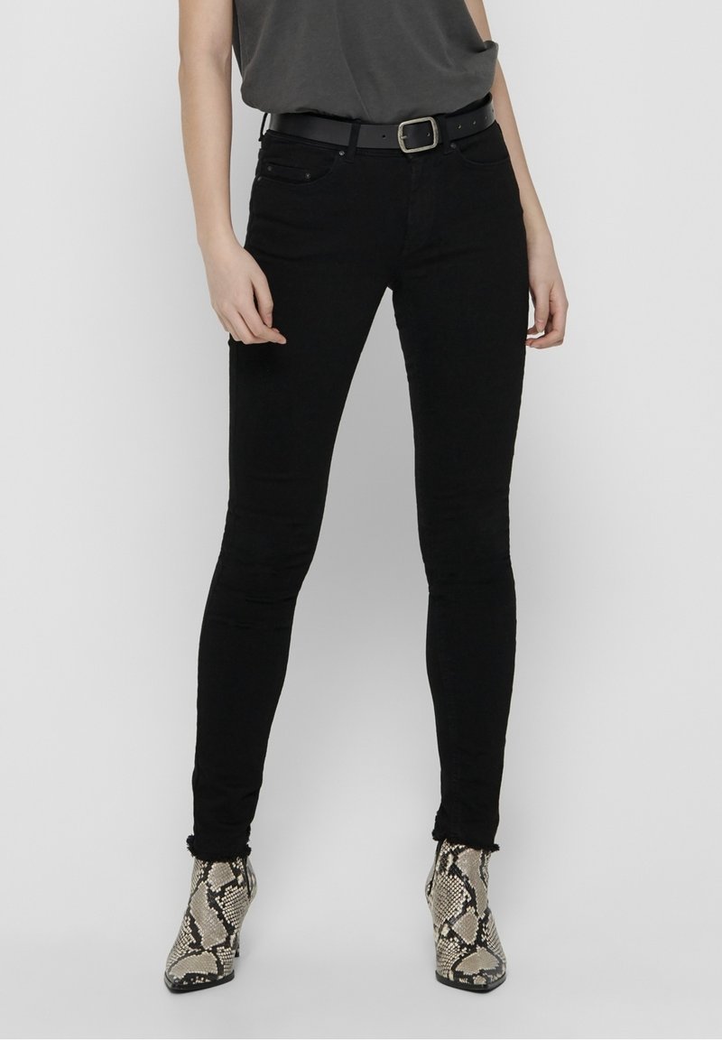 Personne portant un jean slim noir, une ceinture noire avec une boucle argentée, un haut gris foncé et des bottines beiges à imprimé serpent, se tenant devant un fond uni.