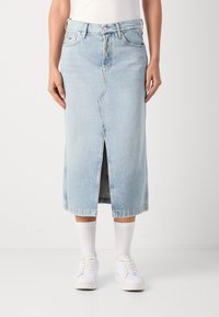 Lys blå denim midi-skort med slisse foran, belteoppheng og klassisk fem-lommers design. Hvite sneakers og sokker er synlige.