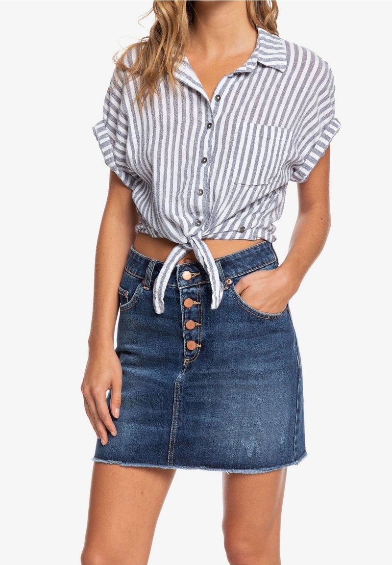Femme portant une chemise rayée à manches courtes nouée et une jupe en jean bleu taille haute avec boutons et poches visibles.