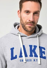 Felpa grigia con un grafico testurizzato blu "LAKE", cordini bianchi e un design casual. Presenta un cappuccio e un tessuto morbido.
