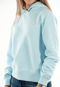 Mujer vestida con un hoodie azul claro de Calvin Klein y jeans azules, mostrando parcialmente el torso y los brazos contra un fondo blanco.