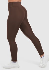 Beine mit braunen Leggings mit hoher Taille, weißen Sportschuhen und weißen, markierten Knöchelsocken, die mit einem Fuß nach vorne auf neutralem Hintergrund posieren.
