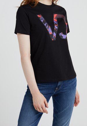Femme portant un t-shirt noir avec de grandes lettres colorées "VJ" et un jean bleu, debout devant un fond clair uni.
