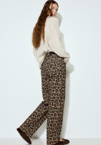 Kobieta o długich falowanych włosach, ubrana w kremowy sweter, szerokie spodnie w panterkę i brązowe futrzane kapcie, stojąca na tle białej ściany.