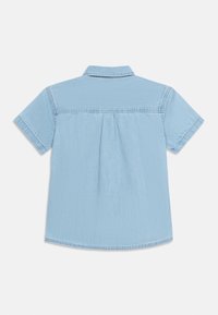 Ανοιχτό γαλάζιο κοντομάνικο τζιν πουκάμισο απλωμένο, με θέα από πίσω που δείχνει ενίσχυση (γιακά) και κεντρική πιέτα κάτω από την ενίσχυση.